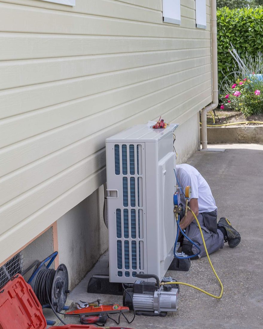 A technician kneeling beside a heat pump outdoor unit on a concrete path against a weatherboard wall, with hoses and a vacuum pump at his feet
